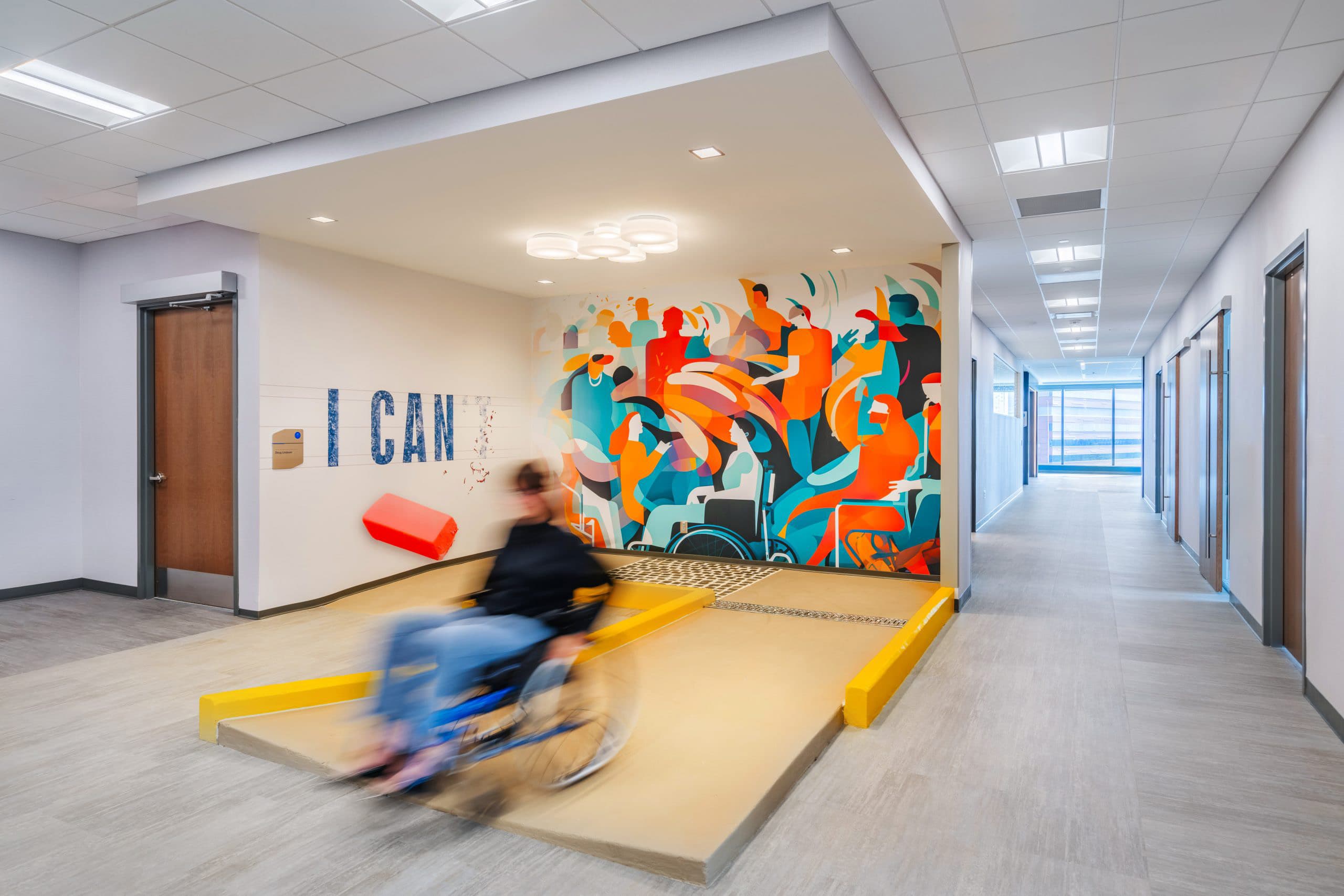 A person in a wheelchair moves along an indoor ramp in a bright hallway, passing by a colorful mural and a wall with large text reading “I CAN.” Several doors and windows line the corridor.