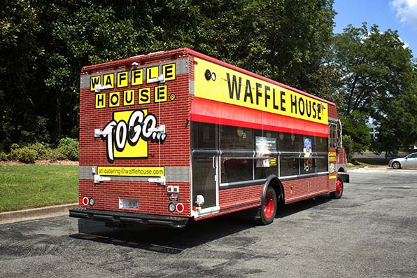A red and yellow Waffle House To Go food truck is parked on a paved lot, surrounded by trees and greenery. The truck has large Waffle House signs and service windows on its side.